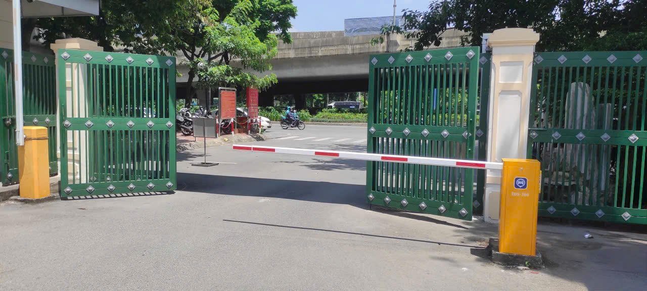 Thanh chắn barie tự động màu trắng đỏ tại cổng ra vào với hai cánh cổng sắt xanh mở rộng